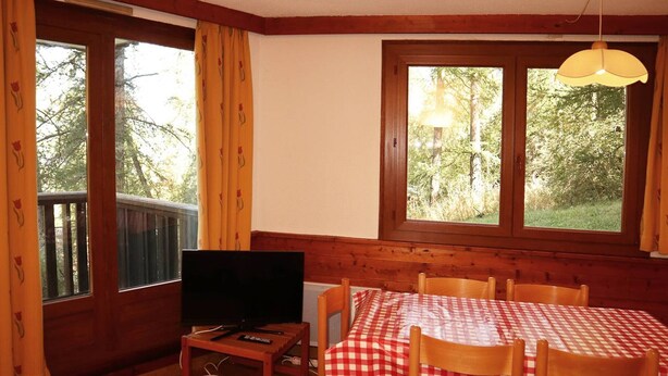 Une pièce avec une table en bois dressée pour deux personnes, un téléviseur posé sur un petit meuble et de grandes fenêtres avec des rideaux.