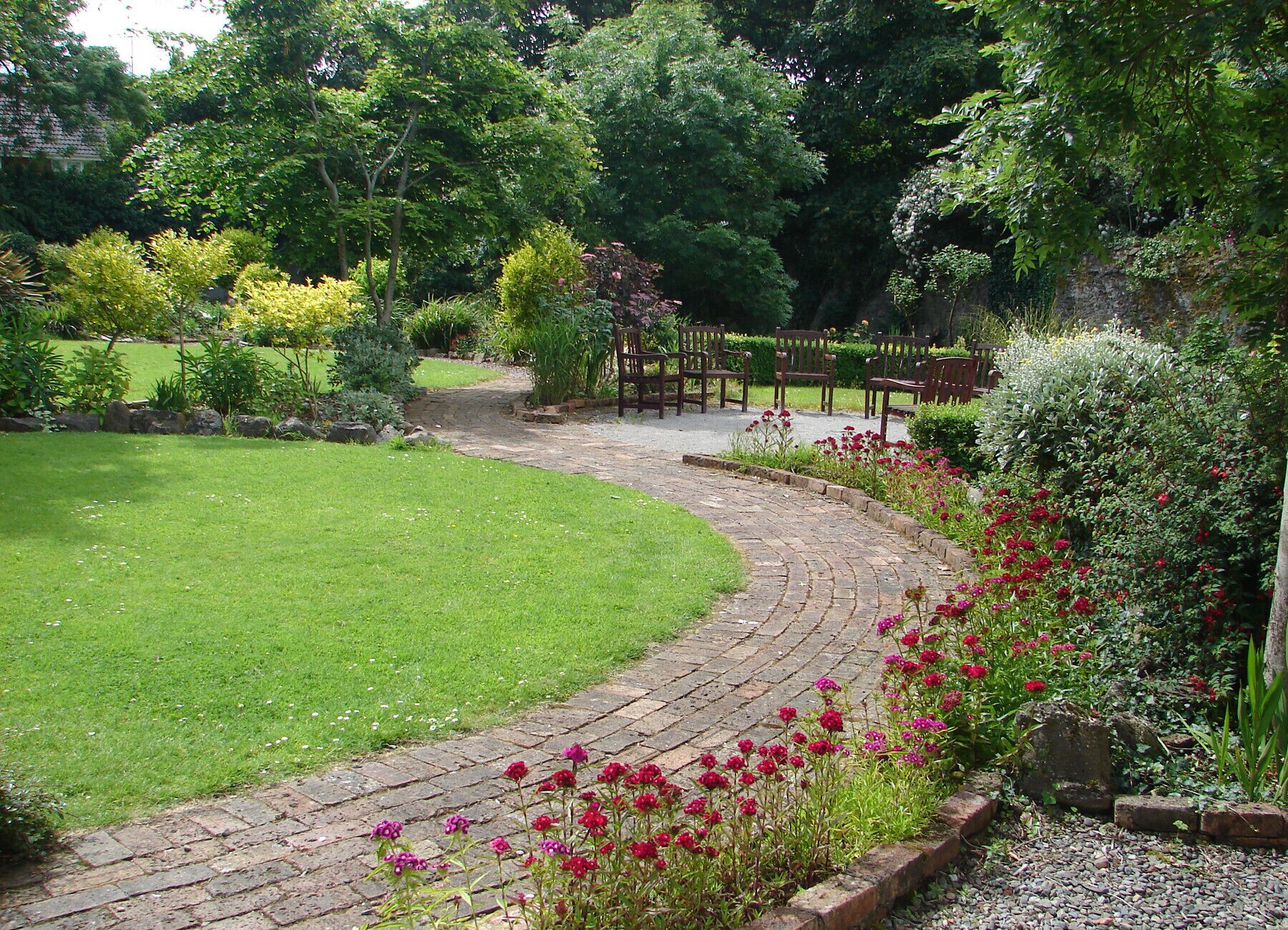 Ein Gartenpfad mit Ziegelbordur, umgeben von üppiger Bepflanzung und blühenden Pflanzen.