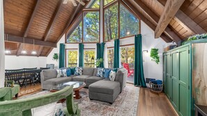 A spacious living room with a large sectional sofa, a coffee table, and a dining area with a table and chairs. The room features a high ceiling with wooden beams, large windows with curtains, and a green cabinet.