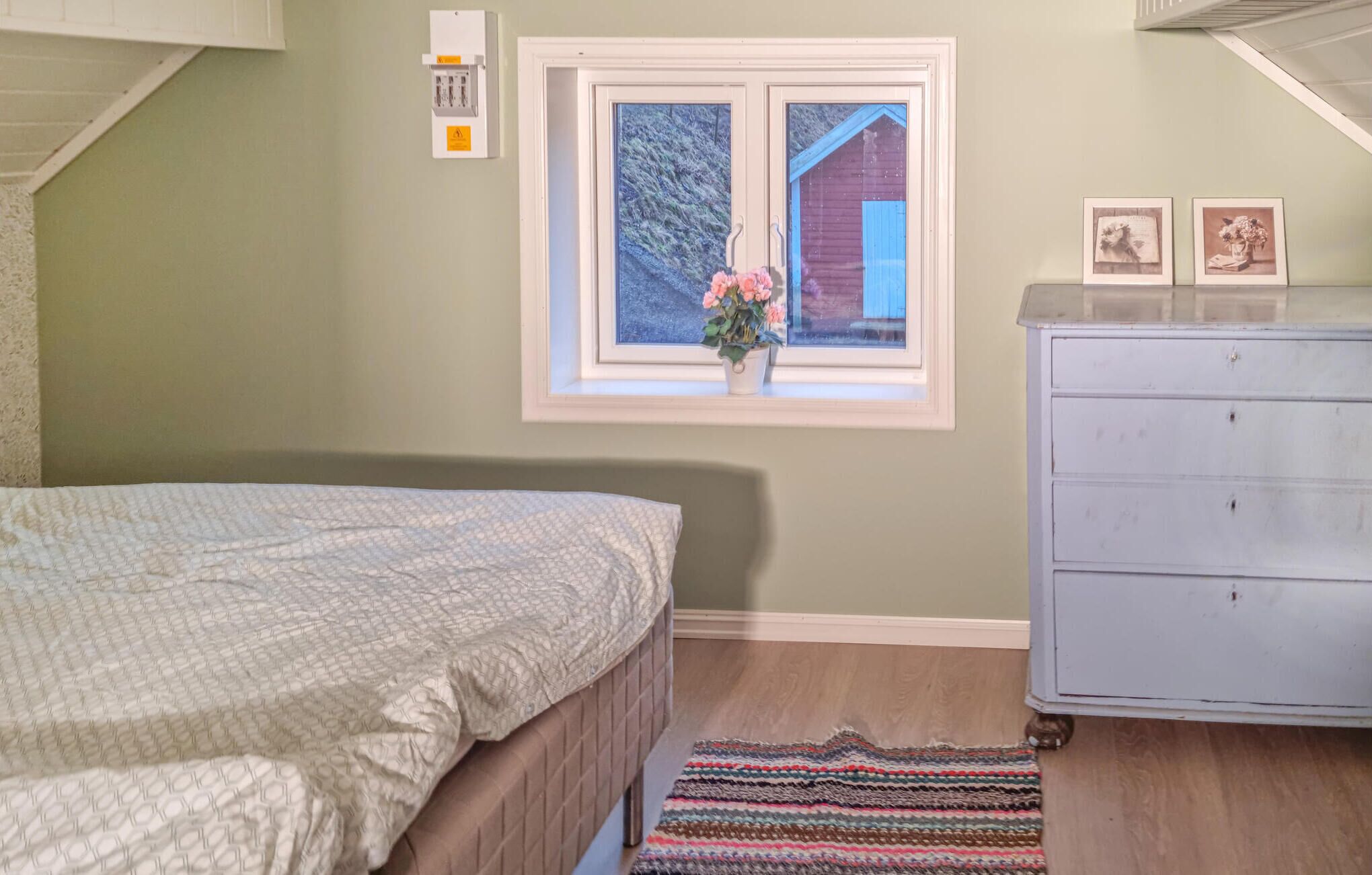 Un dormitori amb un llit, una commode, una finestra que ofereix vista a una casa i un quadre elèctric fixat a la paret.