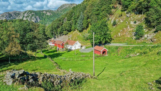 A rural landscape with a stone wall, a red-roofed building, and a white building with a red roof, surrounded by lush greenery and mountains.