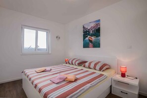 A bedroom with a bed, a window, a painting, and a nightstand with a lamp.