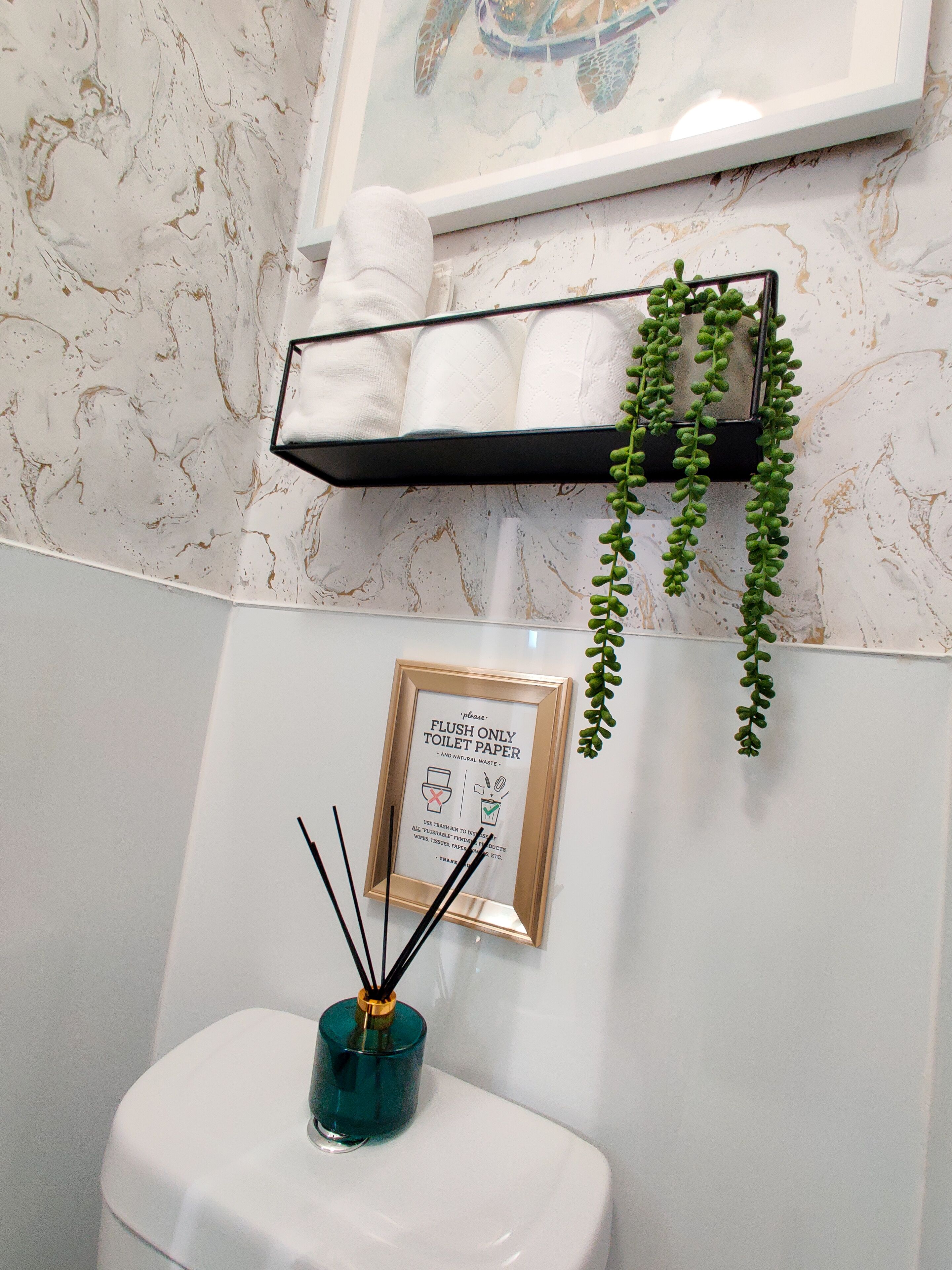 A bathroom with a marble wall, a toilet, a framed sign, a shelf with towels, and a hanging plant.