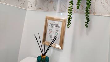 A bathroom with a marble wall, a toilet, a framed sign, a shelf with towels, and a hanging plant.