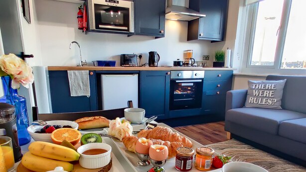 Una cocina moderna con armarios oscuros, un microondas y un horno. Un carrito de desayuno con croissants, frutas y bebidas está sobre la encimera.