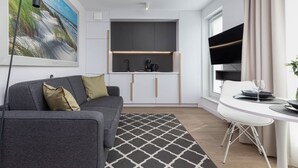 Een moderne woonkamer met een grijze bank, een eettafel met stoelen, een keuken met een zwart aanrecht en een groot raam met gordijnen.