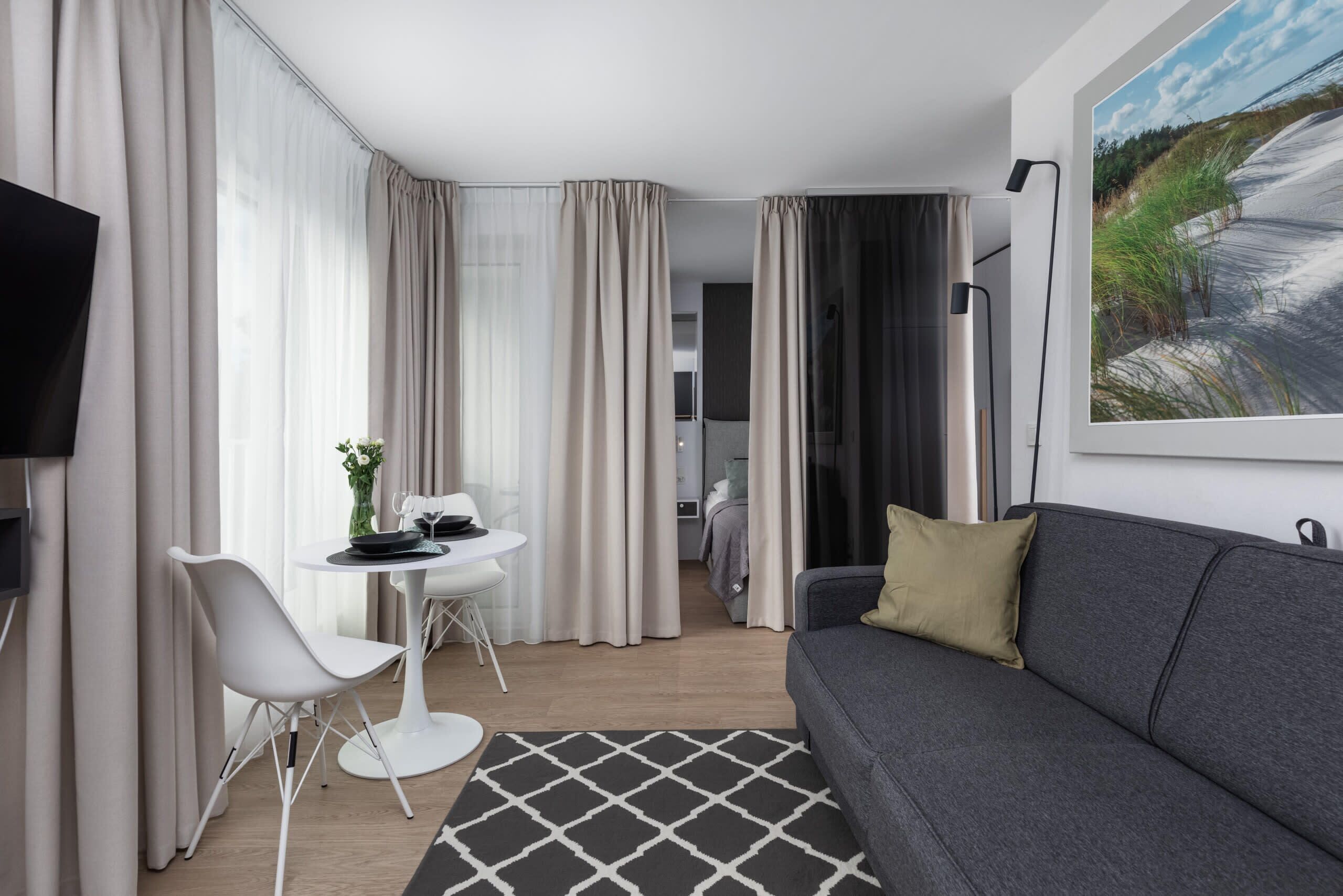Ein modernes Hotelzimmer mit grauem Sofa, einem runden Esstisch mit zwei Stühlen, einem Flachbildfernseher und einem Bild von einer Strandlandschaft.