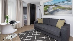 Een moderne woonkamer met een grijze bank, een ronde eettafel en een groot schilderij met een strandthema aan de muur.