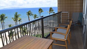 A balcony with a wooden table and chairs, overlooking a beach with palm trees and the ocean.