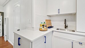 A kitchen with white cabinets, a sink, and a countertop. There is a dish rack with bottles and a wooden tray.