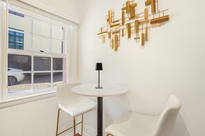 A room with a white round table, two white chairs, a black floor lamp, and a geometric wall art piece.