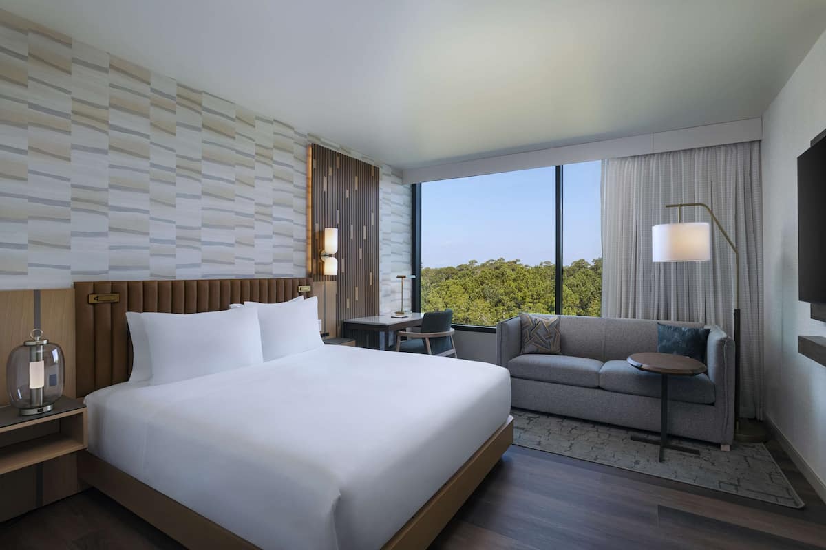 A modern hotel room with a large bed, a sofa, a desk, and a view of the outdoors.