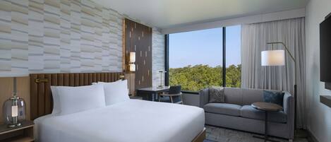 A modern hotel room with a large bed, a sofa, a desk, and a view of the outdoors.