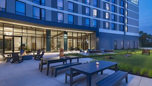 L’extérieur d’un hôtel moderne, avec un espace extérieur bien éclairé où sont disposées des tables et des chaises, de grandes fenêtres et une enseigne « Hyatt Regency » bien visible.