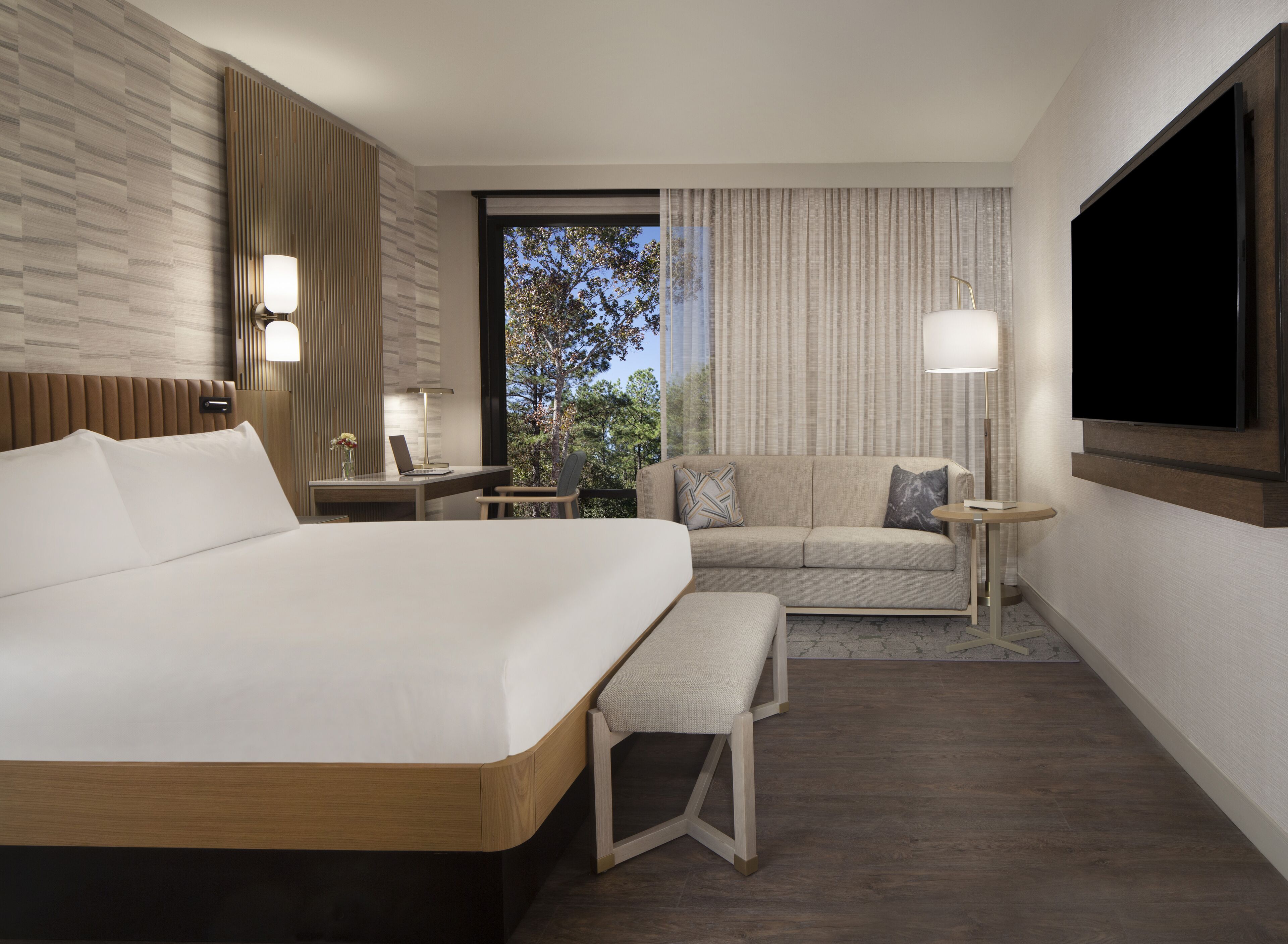 Habitación de hotel con una cama grande, un sofá, un escritorio con lámpara y un televisor montado en la pared.