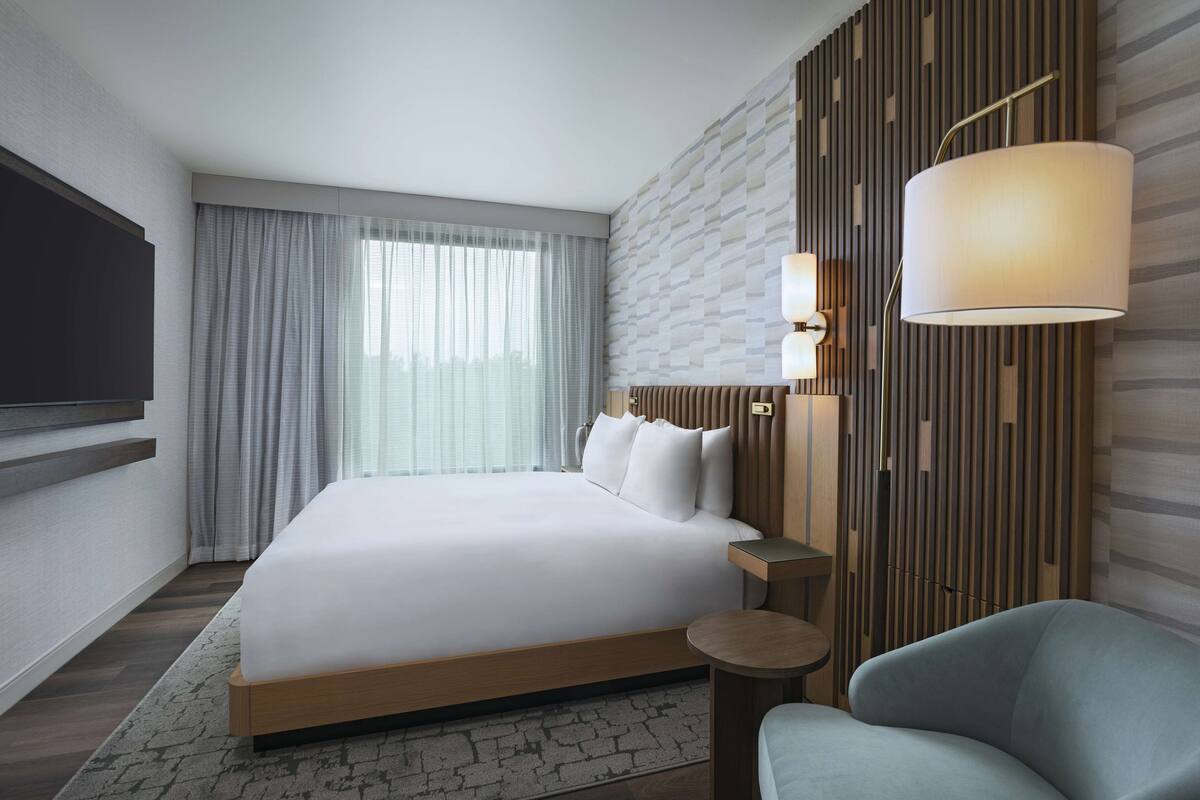 A modern hotel room with a large bed, a flat-screen TV mounted on the wall, a bedside table with a lamp, and a view of the city through the window.