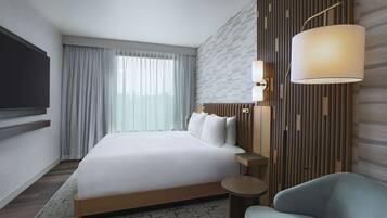 A modern hotel room with a large bed, a flat-screen TV mounted on the wall, a bedside table with a lamp, and a view of the city through the window.