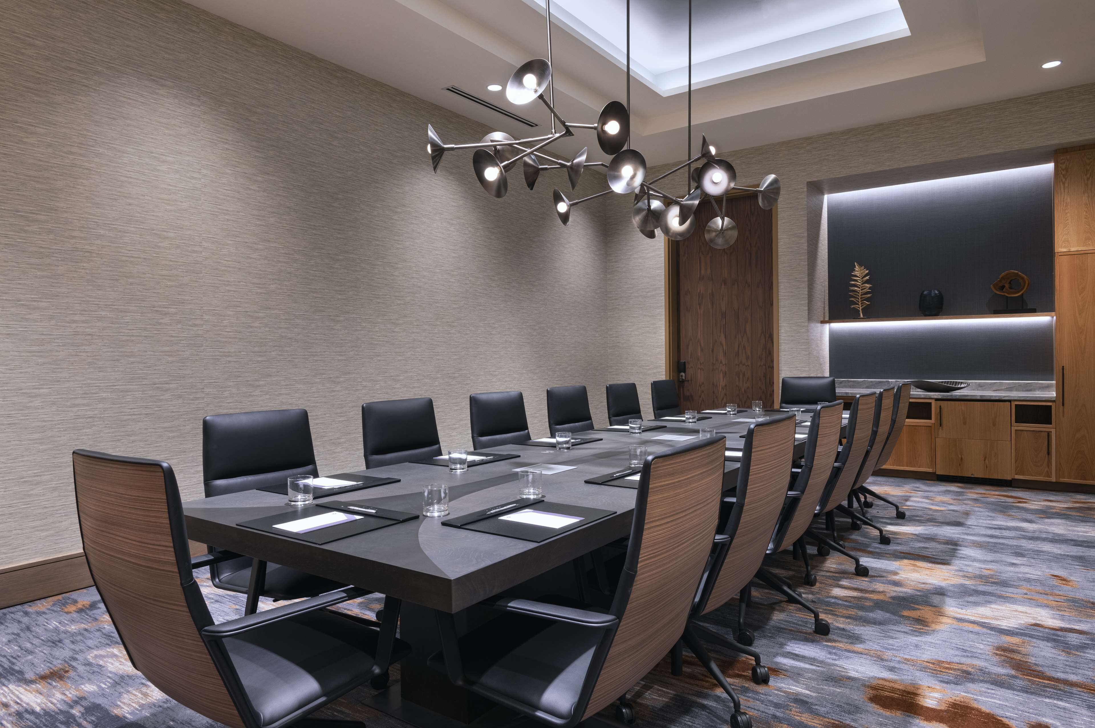 Uma sala de conferências com uma mesa comprida, várias cadeiras, um lustre moderno e um armário de madeira com objetos decorativos.