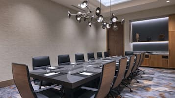 Uma sala de conferências com uma mesa comprida, várias cadeiras, um lustre moderno e um armário de madeira com objetos decorativos.