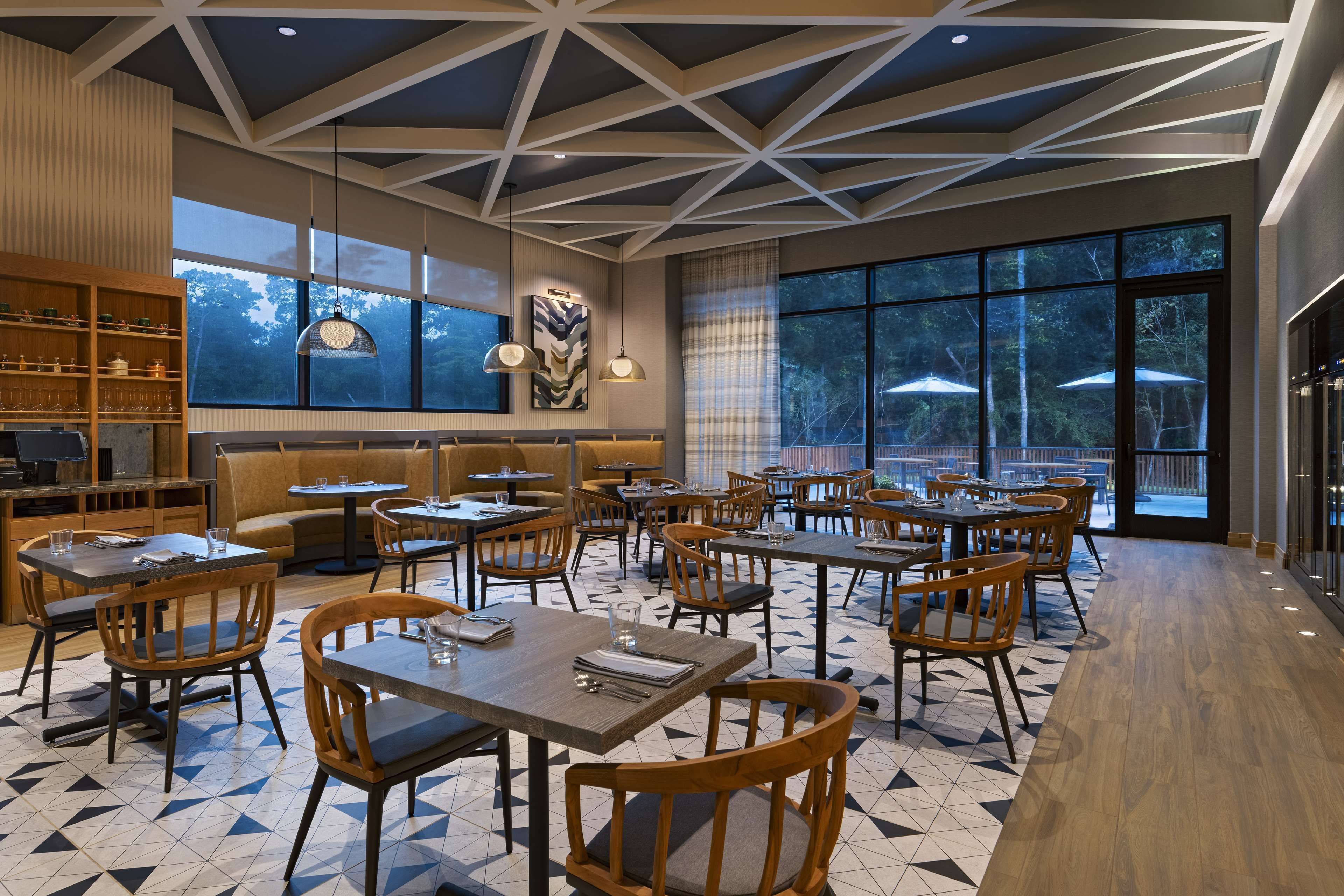 A modern restaurant with geometric ceiling design, large windows, and a mix of wooden and metal furniture.