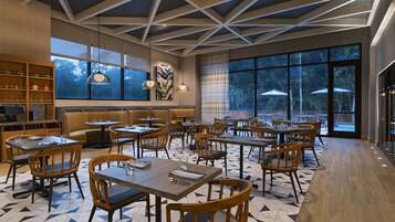 A modern restaurant with geometric ceiling design, large windows, and a mix of wooden and metal furniture.