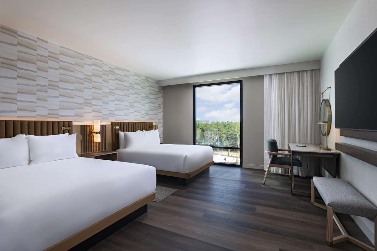 A hotel room with two beds, a desk, a chair, a bench, a TV, and a large window with a view of trees.