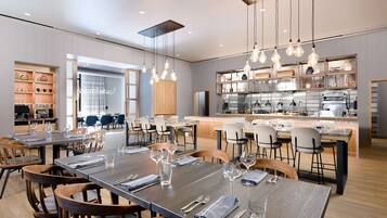 Un comedor moderno con mesas y sillas de madera, una cocina al fondo e iluminación colgante.