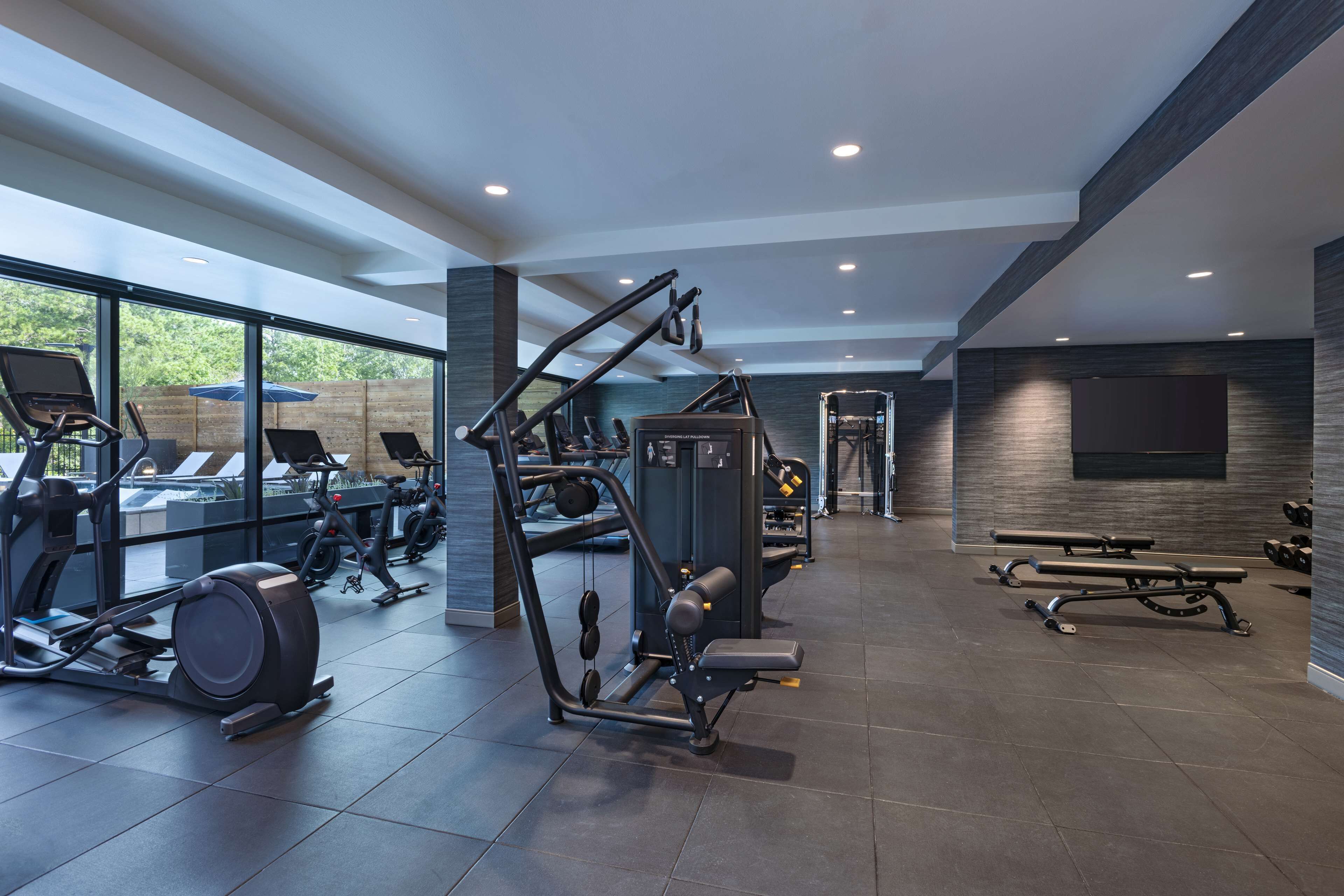 A modern gym with various exercise equipment, large windows, and a bench area.
