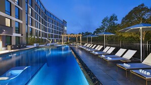 Un espace piscine moderne dans un hôtel, avec une longue piscine rectangulaire, des chaises longues et des parasols.