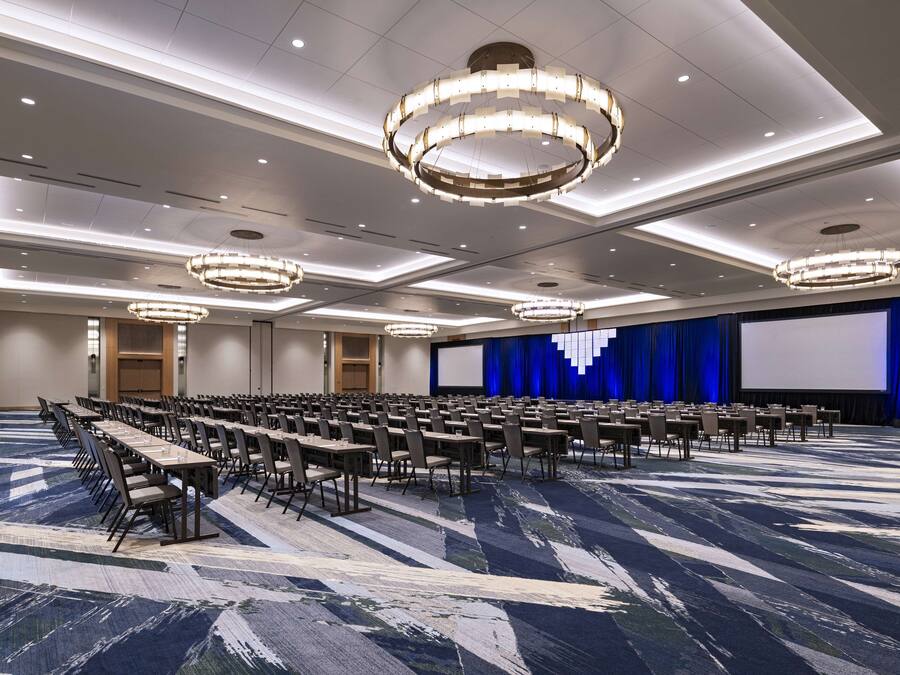 Un salón de conferencias con mesas y sillas en filas, una pantalla grande y un escenario con cortinas.