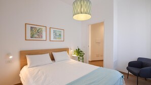 Een slaapkamer met een bed, twee ingelijste kunstwerken, een hanglamp, een kamerplant en een stoel.