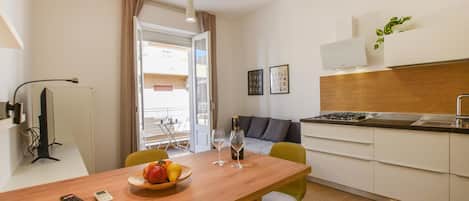 Uma cozinha compacta com área de refeições, uma pequena mesa com frutas e copos de vinho, um sofá e vista através de uma porta aberta.