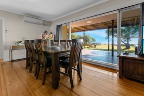 Een eetruimte met een houten tafel en stoelen, een vaas met bloemen en uitzicht op het buitengebied door grote ramen.