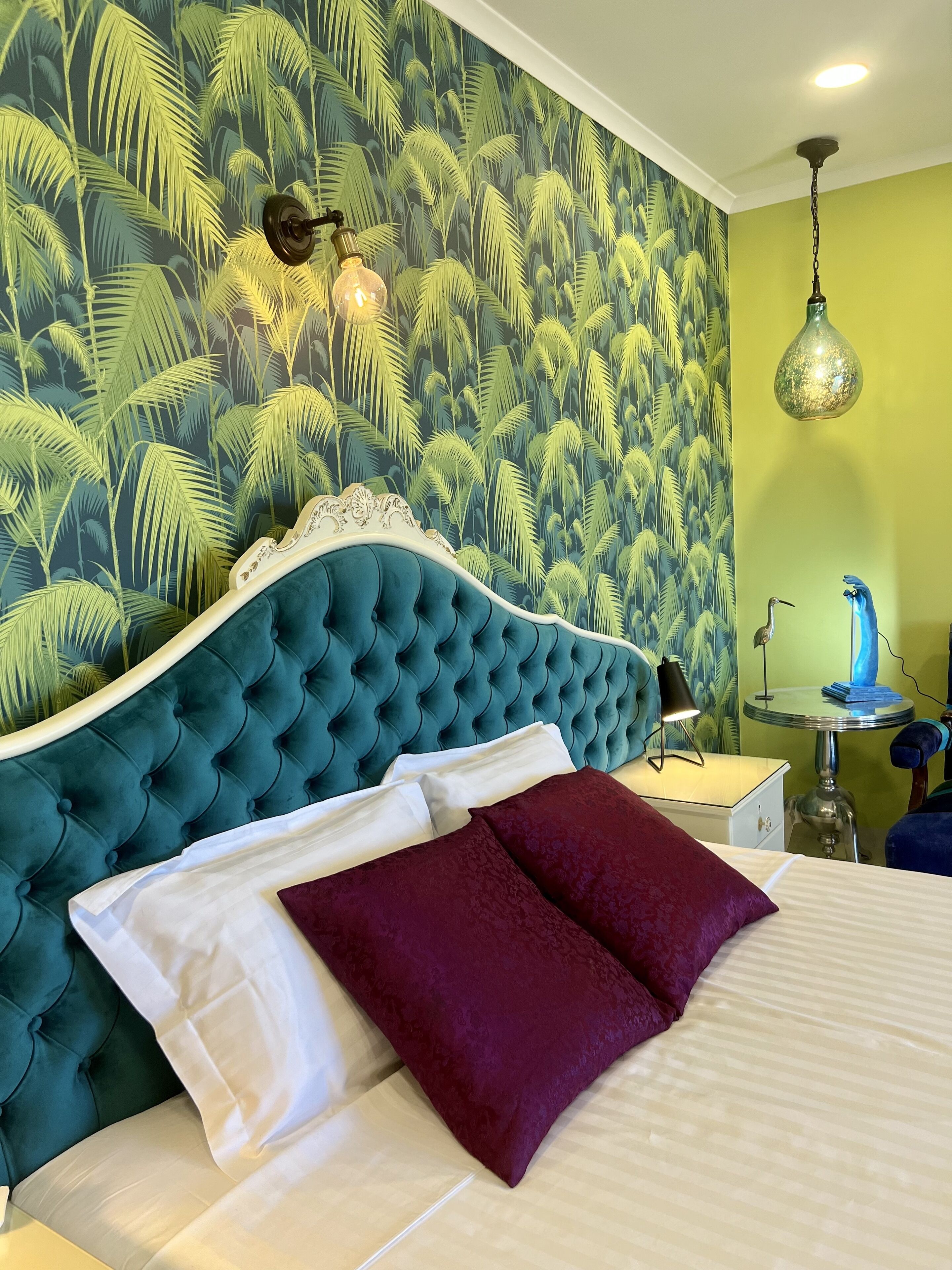 A bedroom with a tufted teal headboard, white and maroon pillows, a bed with a striped bedspread, a wall with a tropical leaf pattern, a hanging light fixture, and a small round table with a lamp.