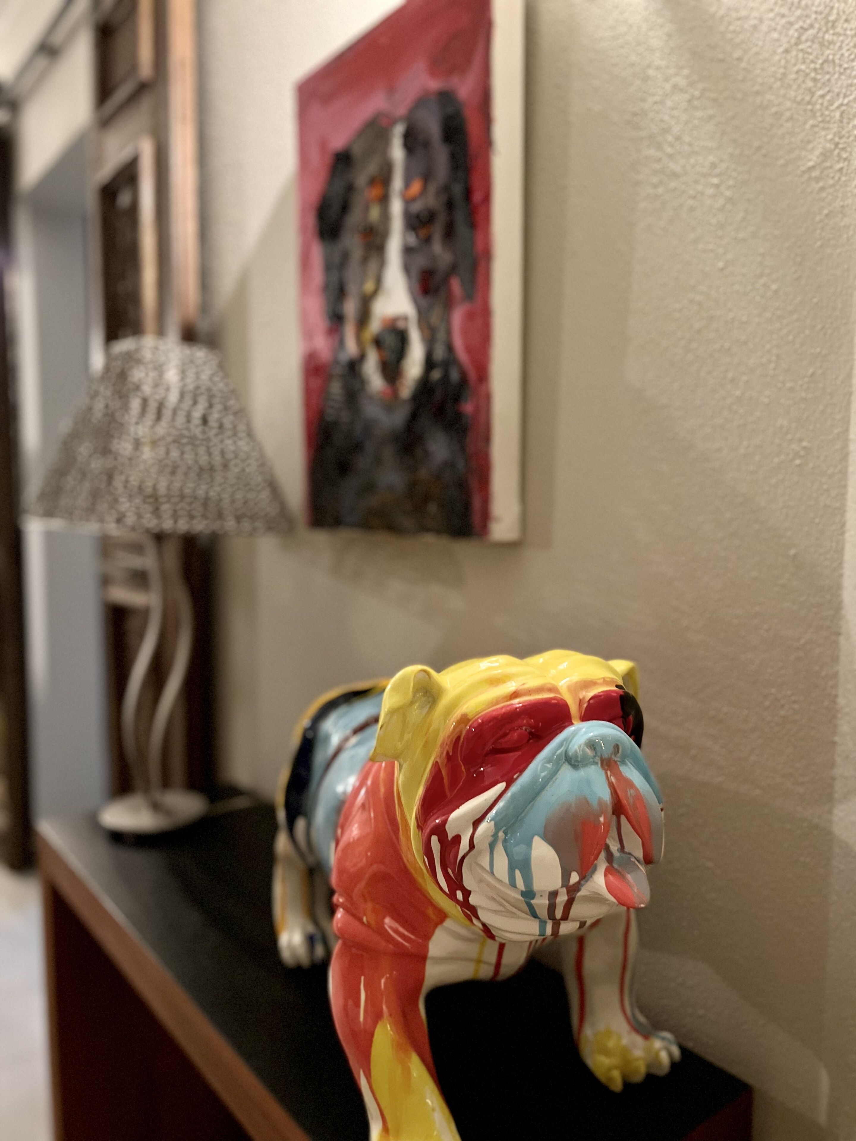 A colorful sculpture of a bulldog on a black surface, a lamp, and a painting of a dog on the wall.