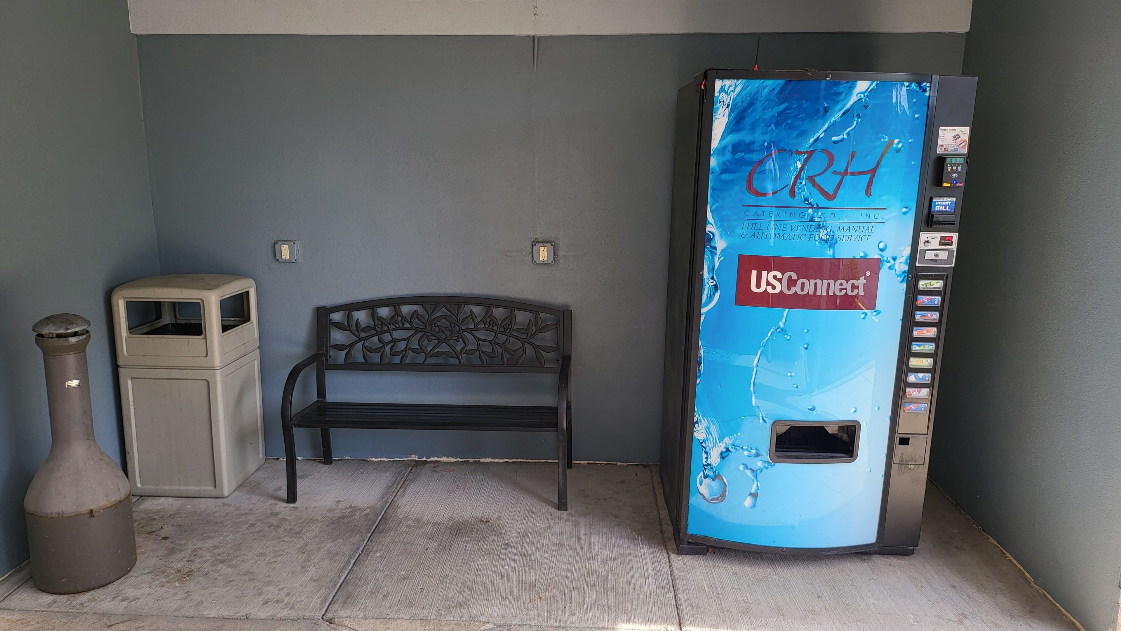 A vending machine with a USConnect logo, a metal bench, and a small trash bin.