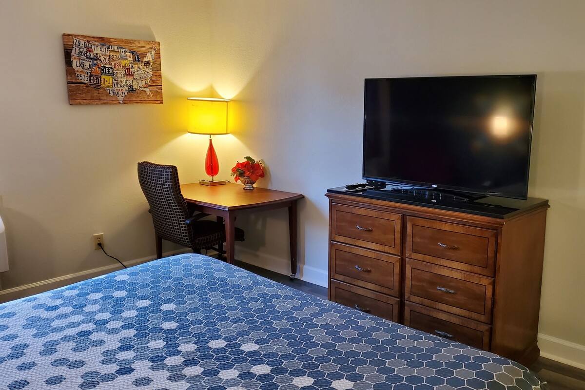 A hotel room with a bed, a desk, a chair, a TV on a wooden cabinet, and a wall-mounted lamp.