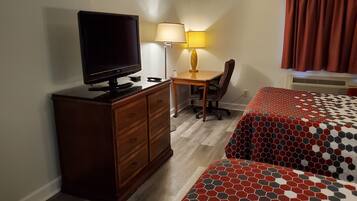 A hotel room with a TV on a dresser, a desk with a lamp, a chair, and a bed with a patterned bedspread.