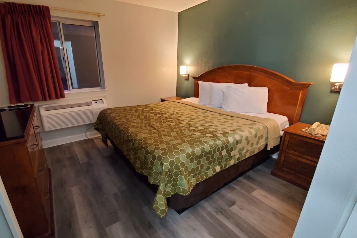 A hotel room with a bed, a nightstand, a telephone, a window with curtains, an air conditioning unit, and a television.