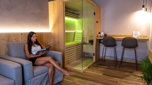 A woman sitting on a grey sofa reading a book in a room with wooden walls and a glass sauna door. - TEVERONE SUITES & WELLNESS - Camera Civetta (Lamosano)