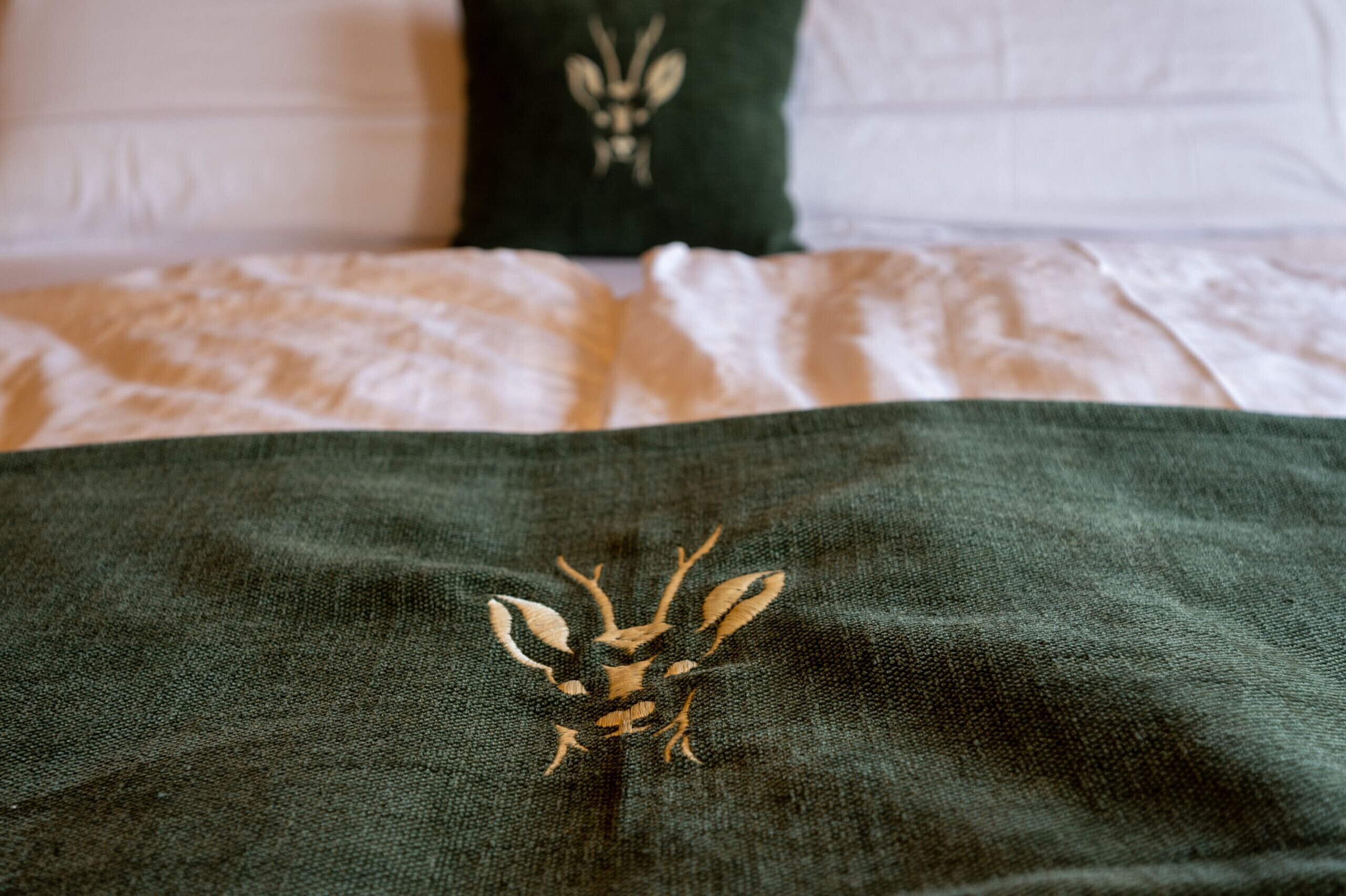 Una cama con un edredón verde con bordado de ciervo, almohadas blancas y una almohada verde con diseño de pájaro.