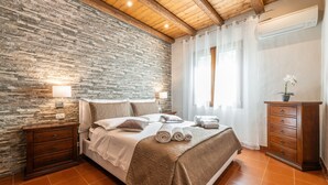 Ein Schlafzimmer mit Steinwand, Holzboden und einem Bett mit brauner Bettdecke, einem Nachttisch, einer Kommode und einem Fenster mit Vorhängen.