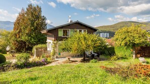 Una casa con techo inclinado, antena parabólica y un jardín con diversas plantas y un banco.