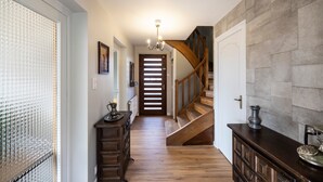 Un pasillo con suelo de madera, una escalera, una araña y una puerta con ventanal.