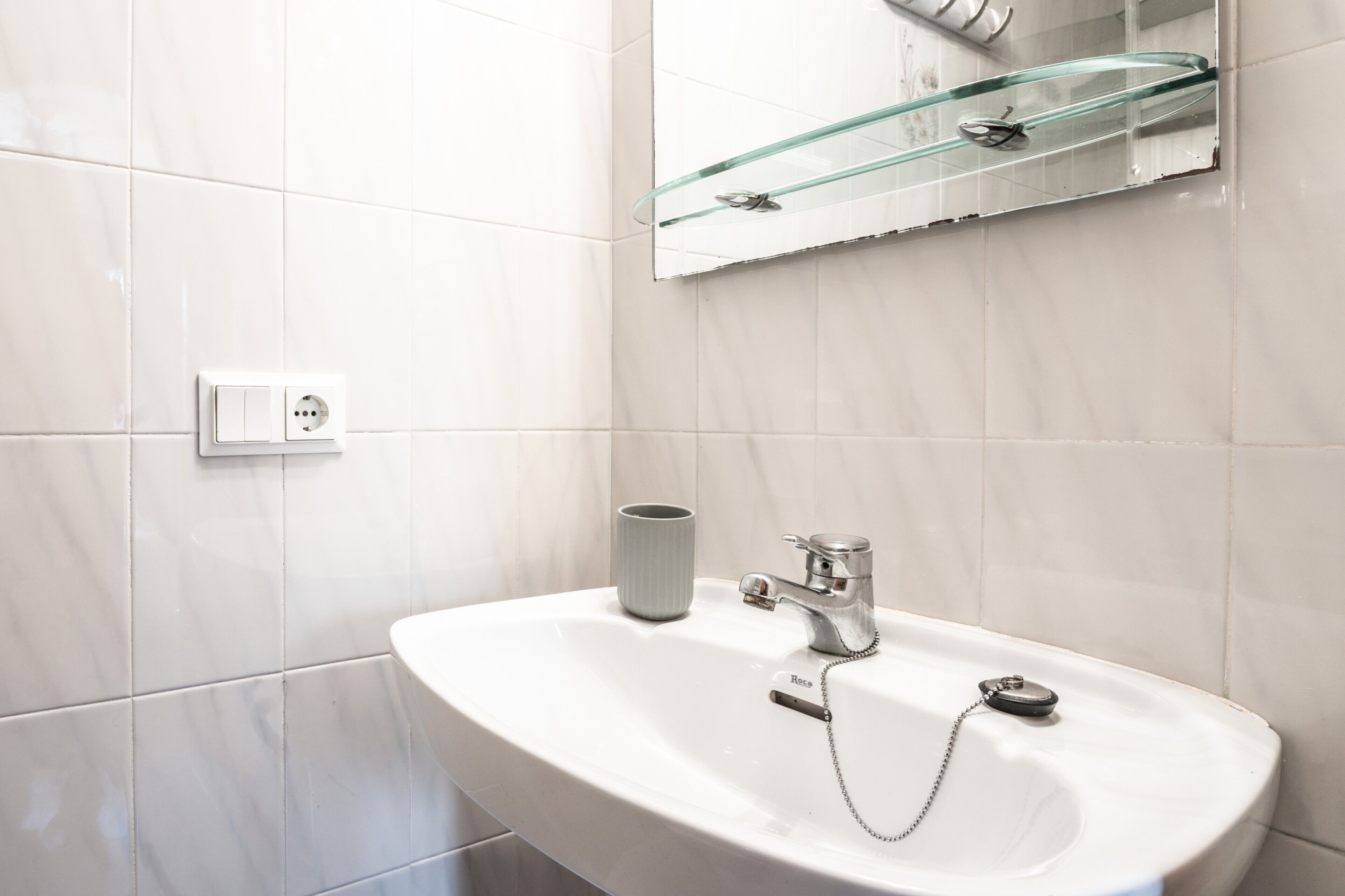 A bathroom with a white sink, a glass shelf above the mirror, and a power outlet on the wall.