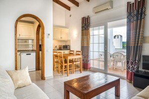 A living room with a wooden coffee table, a sofa, and a TV. There is a kitchen area with a dining table and chairs, and a sliding glass door leading to an outdoor patio.