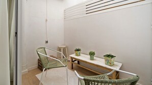 A small, bright room with a white wall, a wooden bench, two wicker chairs, and two potted plants.
