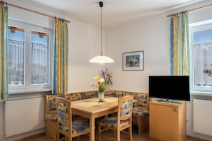 Una habitación con una mesa de comedor, sillas, un televisor sobre un mueble, una ventana con cortinas y un radiador.