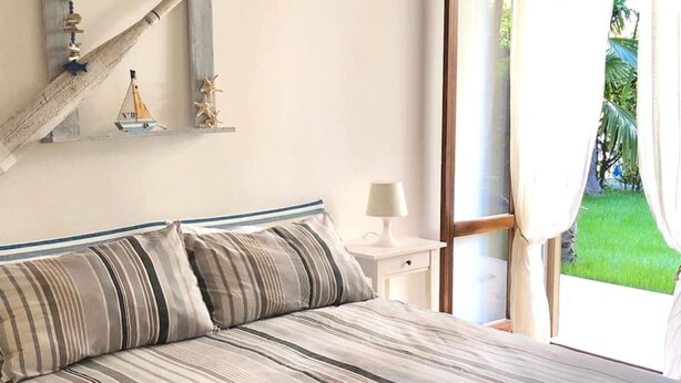 A bedroom with a bed, a nightstand with a lamp, a window with curtains, and a wall with nautical decorations. - Holiday Apartment 'Beba' with Private Garden and Wi-Fi (Lido di Jesolo)