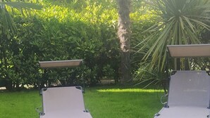 Two lounge chairs in a well-maintained grassy area with a variety of green plants and a palm tree in the background. - Holiday Apartment 'Beba' with Private Garden and Wi-Fi (Lido di Jesolo)